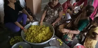 A photograph of a food service being prepared on a concrete floor. In the centre, a large silver metal basin holds yellow rice, and a woman in a brown floral kurta with a dupatta leans over it, reaching in with her hand. A woman in a black top and purple trousers sits on the left with red bangles, beside steel plates holding small round yellow sweets. On the right, a woman in a green blouse and saffron-patterned sari sits with red bangles, and a young child in yellow stands at the edge. Other people in pink and white clothing are partly visible at the top, serving food onto plates.