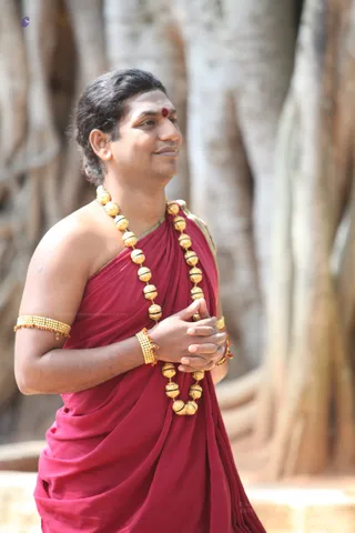A photograph of a man standing outdoors before large banyan tree trunks with aerial roots. He wears a deep red-maroon cloth draped over his left shoulder, leaving the right shoulder and arm bare, and a green-edged border on the drape. He has a long gold-beaded necklace with black accents, gold bead bracelets on his wrists and upper arm, and a red bindi with white sacred ash on his forehead. His hands are pressed together at his chest in anjali mudra as he looks off to the side with a slight smile.