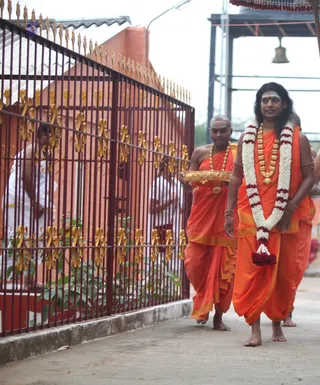 Photograph of a procession on a paved path beside an ornate red metal gate with gold finials and a red-walled building behind. A man in the foreground wears a white garland of flowers with red blossoms hanging at his waist, a gold necklace, bangles and an orange dhoti-style garment, with long swept-back hair and a red tilak mark. Behind him, another man in orange robes and a gold necklace carries a gold tray. A third person in orange is partly visible at the right edge. Two figures in white stand behind the gate, and a bell hangs from a frame at the upper right. a person (appearance-based label: long swept-back hair, ornate gold necklace, floral garland and saffron clothing), walking in procession as the garlanded figure.