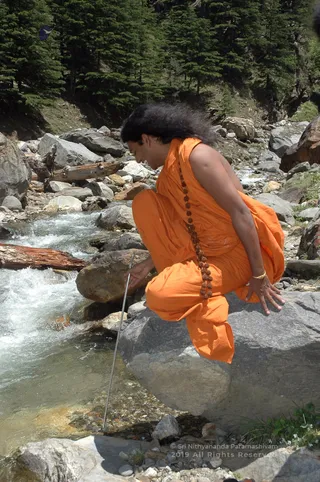A photograph of a person in a flowing saffron-orange sleeveless robe crouching on a large grey boulder beside a fast-running mountain stream. The figure wears a long rudraksha mala draped across the body and a thin gold bangle, with long dark hair swept back. One hand rests on the rock while the other holds a thin metal rod dipped into the rushing water. Behind the stream lie scattered grey boulders, driftwood logs and dense evergreen conifers climbing a steep forested slope.