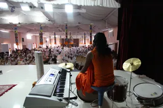 A photograph taken from the stage of a large hall with white draped ceiling and colourful striped hanging pillars. In the foreground, a person seated on a stool at a drum kit faces the audience, wearing a sleeveless saffron-orange top with a saffron-orange cloth draped over the shoulders and long dark hair. A keyboard stands to the left and cymbals and drums are around the stage. Below, rows of seated devotees in white clothes fill the floor, with a few camera tripods and cameramen visible among them. The stage has a dark red curtain on the right.