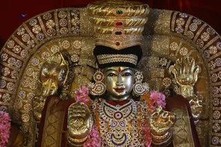 A gilded metal deity murti. The murti wears a tall gold crown set with red, green and blue gemstones, a black velvet band, large jewelled earrings, and a heavily embellished necklace with coloured stones. Its raised hands display lotus-like gestures and it is adorned with pink flower garlands and a red-and-gold silk garment. The overall setting is a temple-style shrine tableau lit with warm light.