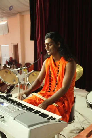 A man with long dark hair, wearing an orange sleeveless garment with a draped orange shawl and a red tilak on his forehead, sits at a white keyboard on a stage while playing drumsticks held in his right hand. A necklace of dark rudraksha beads hangs on his chest. Behind him are two drums labelled with a Western logo, a cymbal, and a dark red curtain. A microphone stand stands nearby, and a few seated people are visible faintly in the background at the left. A person with long hair and orange robes is the visible central figure.