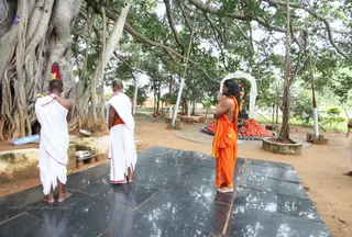 Outdoor photo beneath a large banyan tree with dense aerial roots and hanging vines. Three male devotees stand on a dark stone platform facing a shrine. The figure on the right wears a long saffron-orange robe, wraps the upper body in an orange shawl, has long dark hair and stands with palms pressed together in namaskar. Beside him is a white-robed priest with a shaved head and a white dhoti with a red border, hands folded. Another white-clad man with a shaved head stands at left near a stone platform holding a brass plate with offerings. Garlands of red and orange flowers hang near the tree trunk and a white-arched shrine stands in the background with a mound of red kumkum or offering powder and a deity murti.