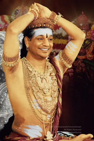 A smiling man seated with both arms raised, hands clasped above his head, bare-chested and adorned with heavy layered gold necklaces, gold bangles and armlets, and a rudraksha crown. He wears a maroon and gold striped cloth draped over his shoulder and a white and vertical bindi-style tilak on his forehead. Behind him are floral garlands in pink, yellow and red and a richly decorated deity murti with a crown. The scene appears to be a formal ceremonial darshan setting. Swamiji (probable): long dark hair, rudraksha crown, ornate gold ceremonial jewelry and saffron-toned adornment are visible.