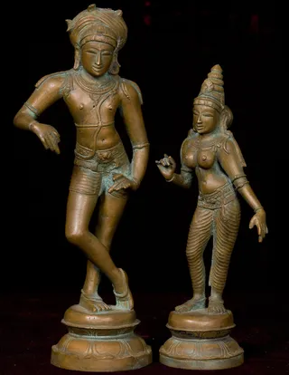 Two bronze statues of a male and female deity stand side by side on lotus-petal bases against a black background. The left figure is a slender male with a tall turban-like headdress, bare torso, a beaded necklace, armlets, and a jeweled waist belt over short dhoti-style lower garments, one leg bent and one hand resting on the hip. The right figure is a female with a tall jeweled crown, bare breasts, a jewelled waist belt and pleated lower garment, one hand raised in a gesture. Patina shows greenish tones on the metal. These are sculptures, not living people, and no a person candidate is visible.