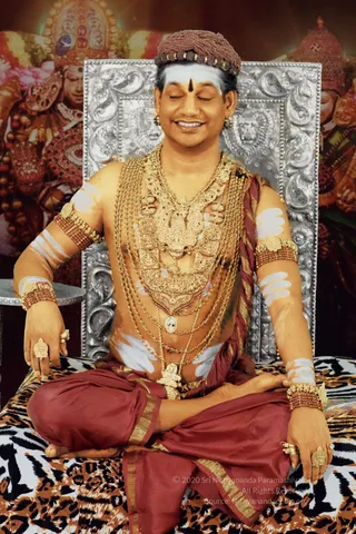 A seated man with closed eyes and a broad smile sits cross-legged on a leopard-print cloth against an ornate silver throne backdrop with lion motifs and a painted deity image behind. He wears a brown beaded crown, a white and vertical red-brown forehead marking, layered gold chains and ornate gold necklaces, beaded brown armbands and gold rings, and a maroon dhoti with gold border. White painted stripes mark his arms and torso, and he is bare-chested. A small framed portrait medallion rests on his chest.