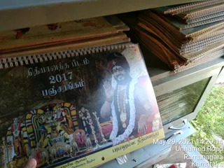 A photograph of a Nithyananda Peetam 2017 Panchangam calendar held in a hand in front of a stack of similar calendars and papers on a metal shelf outdoors. The calendar cover shows a portrait labelled "Paramahamsa Nithyananda" of a bearded man in a red turban and white flower garland with a gold pendant, one hand raised in blessing. Below it are temple deity murtis with garlands and an ornate arch. A date stamp reads May 29, 2021, Unnamed Road, Ramanagara, Karnataka.