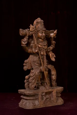 A carved wooden statue of a four-armed deity murti standing on a carved pedestal against a dark background. The figure wears a tall jata-style crown, ornate necklaces and a flowing dhoti with leaf-scroll carving. One upper hand holds a drum-like instrument, another holds a small deer, and a long staff or trident-like rod rises beside the body. The lower hand is open in a gesture. The base has relief carving of a small figure. The wood is a warm dark brown with visible grain.
