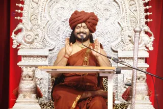 A seated man in saffron-orange robes and a rust-coloured turban sits on an ornate silver throne with lion-head armrests, before a red curtain. He has long dark hair, a rudraksha-style beaded mala and a gold necklace, and raises both palms in greeting or blessing while smiling with eyes closed. A wooden lectern and microphone stand in front of him. Swamiji (probable): the saffron robes, turban, long hair and gold necklace fit the description, and he is seated on a throne as the central speaker.