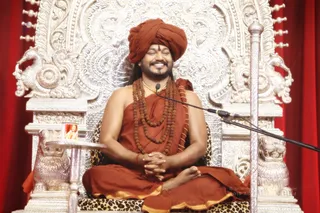 A man seated cross-legged on an ornate silver throne with intricate repoussé carvings and lion-head armrests, against red draped curtains. He wears a rust-orange turban, orange-saffron robes with gold trim, multiple rudraksha malas and a gold necklace, and holds his hands folded in his lap while smiling with eyes closed. A microphone on a stand is at his side, and a small framed portrait sits on a silver side table. Swamiji (probable): long hair visible, orange turban and saffron robes with rudraksha malas and gold ornaments; seated in a throne as the speaker. A leopard-print cushion lies on the throne seat. Ashram appears at the lower right.