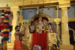 A priest in orange-saffron dhoti and shawl, with a bare torso and rudraksha mala, stands at left pouring or lifting a vessel over a golden deity setup. Three deity murtis sit on a golden ratha-style vahana or throne with red cloth draped over its base: a central figure in a crown with a green and gold garment, flanked by two figures in red and gold-trimmed garments with floral garlands. Hanging bells, gold pillars, and tassels in red, green, yellow and purple decorate the temple hall. A small metal pot and a seated attendant appear at lower right. The scene suggests abhishekam or puja in a temple shrine.