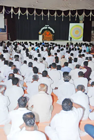 Photograph from behind a large hall audience seated cross-legged on the floor, most wearing white shirts and white dhotis, with a few in other colours. On the stage at the back, a figure in orange robes sits on an ornate brown throne-like chair behind a small white podium with a flower arrangement. Above the stage, a banner reads NSP and Nithyananda Smaranai in Tamil script. To the right of the stage, a large backdrop shows concentric green, yellow and orange circles, and a video camera on a tripod stands near the stage. The hall has maroon and white swagged drapes and ceiling fans overhead.