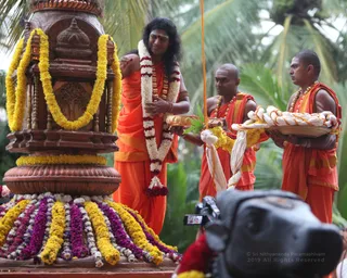 Outdoor ritual photograph beside palm trees. A long-haired man in orange robes with a white and red flower garland stands at a carved wooden pillar-like shrine wreathed in yellow and purple-white flower garlands. Two shaved-headed attendants in orange robes and rudraksha malas stand behind him; one holds a brass plate with leaves, the other a tray of white and yellow cloth. A video camera is in the foreground at right. Appearance cues are moderate, so classified as in_person.