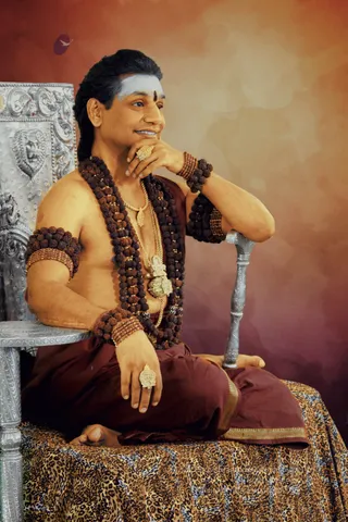 A seated man in a silver-embossed throne chair with lion motifs, draped in a leopard-print cloth. He is bare-chested with a long dark rudraksha mala across his chest and multiple rudraksha armlets and bracelets, gold chain necklaces with a pendant, and gold rings. He wears a maroon dhoti with gold border, has white forehead markings with a central dark tilak, and rests his chin on one hand while looking off to the side against a warm orange and red backdrop.
