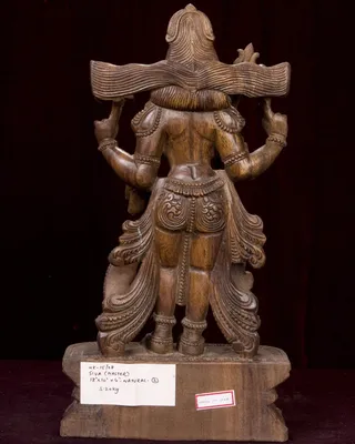 Rear view of a carved wooden statue of Lord Shiva standing on a rectangular wooden plinth against a dark maroon backdrop. The deity has multiple arms raised, holding objects overhead, with an elaborate high topknot and flowing carved drapery, jewelled armbands and a decorated waist band. A paper label on the plinth reads WK-15/68 SIVA (MASTER) 18x10x6 NATURAL and 5.20 kg, and a small red-bordered tag reads MADE IN INDIA. No people are visible.