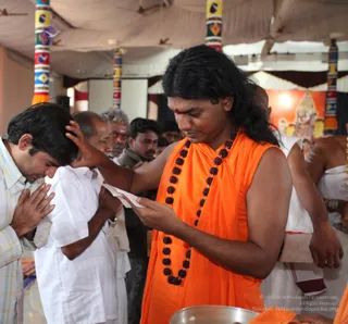 A photo of a ritual blessing in a tented hall with colourful striped pillars and a deity print on a red backdrop. A long-haired man in a saffron-orange sleeveless wrap with a long rudraksha mala and a red tilak stands center-right, reading a folded paper while placing his right hand on the head of a bowed man in a light checked shirt with hands joined in namaskar. Beside them an older man in white shirt and dhoti stands, and other devotees watch in the background. A metal bowl sits at the lower edge. Probable Swamiji is the central figure: long swept-back hair, saffron wrap and rudraksha mala. Count: about six visible people.