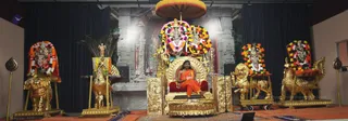 Wide photo of a satsang stage with a gilded throne at centre, draped in red velvet and topped by a large yellow-and-red umbrella. A man in saffron-orange robes with long dark hair sits on the throne, a microphone stand before him, appearing to address an audience. Behind him, deity murtis are garlanded with bright multicoloured flower garlands on a grey stone-textured backdrop. Flanking the stage are gilded lion and horse-shaped seats with floral garlands and peacock-feather fans, and a pair of ornate chairs with garlanded deity figures. Dark blue curtains frame the set. Printed caption reads Sri Nithyananda Paramashivam at the lower right.