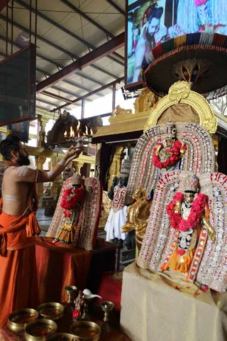 A photograph inside a golden-pillared temple hall of a priest bare-chested with white sacred markings on his arms and chest, wearing an orange cloth wrapped around his waist and shoulder, holding a lit multi-flame oil lamp aloft toward two richly decorated deity murtis. The murtis are dressed in embroidered silver-and-red garments, with red and pink flower garlands and gold crowns, seated before ornate gold arches and a decorated umbrella canopy. In the background, a white-clad figure stands near a black stone sculpture, and brass bowls with offerings sit on a red cloth at the lower left. A large screen above shows a video of people. The scene depicts an aarti performed before the deities.