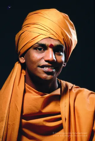 Head-and-shoulders studio portrait of a smiling man against a plain black background. He wears an orange-saffron turban and an orange-saffron shawl draped over the shoulders, with a red kumkum mark on the forehead and a dark mala visible at the neck. The , 2019 .