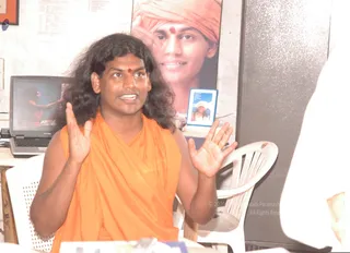A man with long, shoulder-length dark hair and a red bindi sits on a white plastic chair, speaking and gesturing with both hands raised. He wears an orange saffron wrap draped over his shoulders. Behind him a large printed portrait of a smiling figure in an orange head wrap and red forehead mark hangs on a wall, and a laptop on a table shows a screen with a figure in orange. A small framed card stands on the table. A partially visible person in white sits at the right edge.