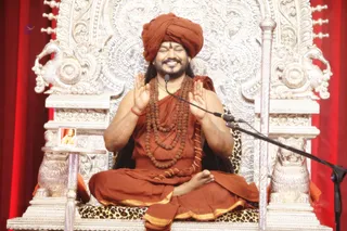 A seated man in an orange-saffron turban and matching saffron robes sits cross-legged on an ornate silver throne with carved lion and peacock motifs and a leopard-print cushion. He wears multiple rudraksha malas and a gold chain, and holds his palms together in front of his chest with a smile. A microphone stands before him, and a small framed portrait is visible at left. Red curtains form the backdrop. Swamiji (probable): the saffron turban, long swept-back hair, rudraksha malas and saffron robes match the candidate's distinctive presentation, so the figure is labelled as probable in person.