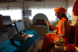 A man in saffron-orange robes and an orange turban sits on an ornate red and gold throne, raising one hand in a gesture while speaking. He wears a rudraksha mala and a long orange shawl. Beside him is a golden lion-shaped figure at the base of the throne. In front of him on a teal draped table are a monitor and a laptop stacked on books, showing a video call with an image on screen. A pale green armchair with a red cushion, potted palms and white curtains fill the room, with a lighting stand behind.