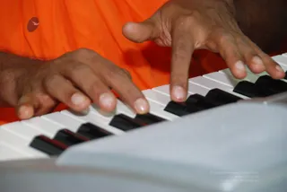 Close-up photograph of a pair of hands playing a white electronic keyboard with black keys. The person wears orange-saffron cloth visible at the edge of the frame. A small red-and-white bird logo sits in the upper left. Only hands and orange clothing are visible, with no face shown.
