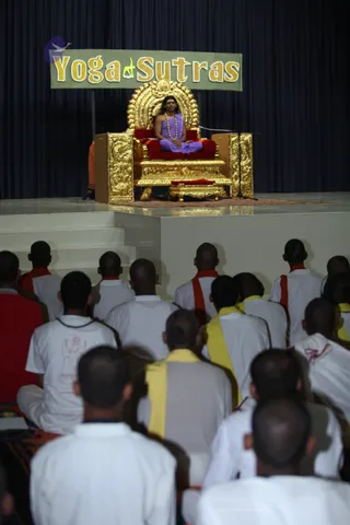 A photograph of a satsang hall with a raised stage. On stage, a man with long dark hair sits cross-legged on an ornate gold-and-red throne under a banner reading "Yoga Sutras". He wears a lavender wrap over bare shoulders and a long beaded necklace, and a microphone stands beside the throne. In the foreground, rows of seated devotees face the stage from behind; most wear white clothes with red or yellow shawls, and several have shaved heads. Dark blue drapery covers the back wall.