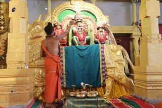 A photograph inside a gilded temple hall showing a priest in an orange dhoti and orange shawl with a bare upper body, standing with his back to the camera and applying a mark or touching the head of a deity murti. The murti is seated on a gilded peetha or throne with a golden lion-like carved base, wearing a crown, pink and red flower garlands and a green backdrop. Two smaller seated figures flank it, also garlanded. In front, a teal and purple cloth covers the platform with brass bowls, a kalasha-like vessel and offerings on a striped mat. The scene is an abhishekam or puja setting. Golden pillars frame the scene.
