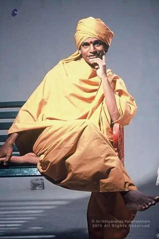 A photograph of a single seated man wearing a saffron-yellow turban and a flowing saffron-yellow robe wrapped around his shoulders and body. He sits on a green slatted bench with a red metal armrest, one bare foot raised, and rests one hand against his chin while gazing off to the side. A small red bindi is on his forehead. A translucent plastic cup sits on the grey floor nearby. The background is a plain grey wall. A copyright notice is printed over the lower right corner.