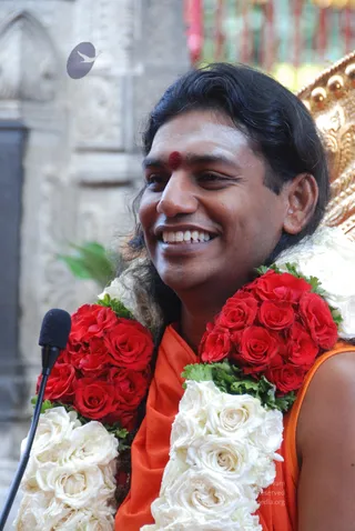 Close photo of a smiling man in an orange saffron garment with a red bindi on his forehead and long swept-back dark hair. He wears heavy garlands of red roses and white roses over his shoulders and sits beside a black microphone on a stand. Behind him is a gold-toned ornate chair and a stone wall with carved details. The swami's appearance matches probable Swamiji cues: swept-back long hair, saffron clothing, and ornamented seating, with the page stating he is present at this event.