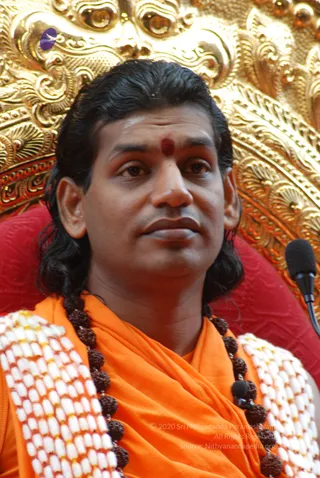 Close photo of a man seated in front of an ornate gold-embossed backdrop with a red cushioned chair. He has long dark hair swept back, a red kumkum dot on his forehead, and wears an orange saffron wrap with a dense rudraksha mala and a white beaded shoulder garland. A black microphone stands at the right edge.