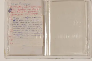 A photograph of an open handwritten puja manual inside a clear plastic sleeve, photographed flat on a light background. The left page holds red and blue ink handwriting in Tamil script, with a small loose slip of paper in blue ink laid over the lower part of the page. The right page is blank and cloudy under the plastic. No people are visible.
