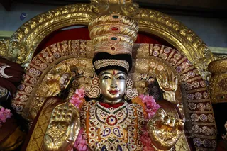 A gilded deity murti of the goddess Meenakshi, seen close up against a richly embellished golden arch. She wears a tall jewelled golden crown set with red, green and white stones, a black velvet band beneath it, and large round gold earrings. Her chest is covered by an elaborate jewelled necklace and bodice in red, green and white stones with ornamental ornaments. Her raised gilded hands hold ceremonial items, and pink flower garlands drape over her shoulders. Behind her, red velvet and gold filigree panels frame the image, with a crescent moon motif at the left edge. No people are visible.