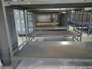 Interior photograph of an empty dormitory-style room fitted with rows of grey metal bunk beds and bare metal slats, one bed topped with a thin mattress. Lockers stand along the left wall and the floor is pale tiled. A timestamp in the lower right reads 30-May-2021 5:40:00 PM. No people are visible.