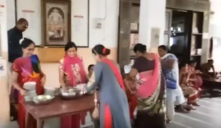 A blurry video still of an indoor hall where several women in salwar kameez and sarees serve food. A woman in a red and blue dupatta stands at a wooden table ladling from steel bowls and pots, while others in red and pink attire carry plates. Seated women and a seated man in a dhoti-like wrap sit along the right side near a doorway. A framed devotional image of a deity hangs on the wall behind them. The people visible total about eight, and no clear a person candidate is identifiable from appearance.