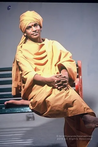 A seated man in a yellow-orange turban and matching yellow-orange draped robe sits on a green slatted bench with a red-painted backrest against a plain grey backdrop. He rests his clasped hands on his lap, has a red dot on his forehead, and his bare feet are visible, one leg extended forward. The image has a studio portrait feel with a pale grey background. Swamiji (probable) appears as the central figure, identified by a turban, saffron-toned robe and forehead mark. A small logo appears at top left, and a 2019 ' at lower right