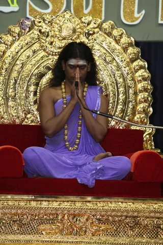 A seated man with long dark hair, a white forehead marking and a red bindi sits cross-legged on a red velvet seat in front of an ornate gold throne with a large gold arched backrest. He wears a lavender-violet draped cloth and a long necklace of gold beads, with palms pressed together in namaskar and eyes closed. A microphone on a flexible stand is angled near his hands. The background shows gold lettering on a grey panel. a person: long swept hair, gold ceremonial necklace and ornate gold throne are the qualifying cues; the figure is in a seated devotional posture during a talk setting.
