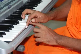 Close-up photo of a person in an orange saffron robe playing a white electronic keyboard with black keys. Only the hands and forearms are visible, with fingers pressed on the white keys while the body remains cropped out of frame. The keyboard sits on a grey stand, and a faint blue logo appears on the keyboard's top-left panel.