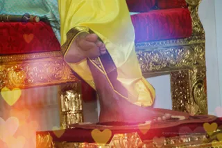 Close-up photograph of a seated person's bare foot resting on a red cushioned ornate golden throne platform. The person wears a yellow silk dhoti with a gold border and a dark maroon trim. Behind the foot, a red velvet cushioned seat with gold-carved armrest panels and a golden carved leg. A bright blue and white patterned fabric is visible at the upper left. Pastel heart-shaped overlays float across the lower left and right corners, and a small purple rudraksha-like ornament appears near the top left. Only the foot and lower leg of one person are visible, so the figure cannot be identified from the frame.