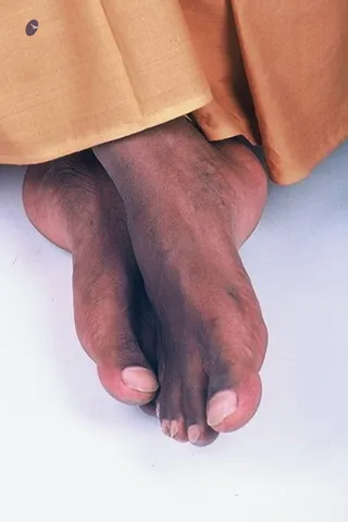 Close-up photograph of a pair of bare feet resting on a plain white surface, with the legs disappearing under the hem of orange-saffron cloth. The feet are crossed and show visible toenails and fine hair on the skin. The orange garment is draped at the top of the frame. The image is cropped tightly to the feet, with no face or other body parts visible.