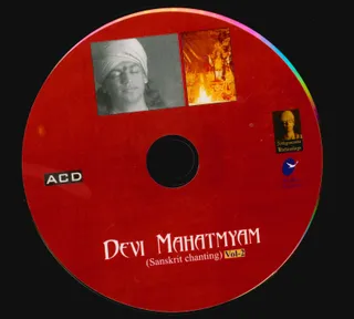 A red compact disc label titled Devi Mahatmyam (Sanskrit chanting) Vol-2. Two small photographs sit at the top of the label: on the left, a black-and-white portrait of a man with closed eyes, a tilak mark on his forehead and a white turban; on the right, a deity murti with a flame in front of it. A small a person Kirtanalaya logo and a Life Bliss Foundation bird emblem appear on the right, and an ACD marking on the left. Appearance-based cues on the portrait are insufficient for a confident identification, so the label is recorded as a depiction with uncertain identity.