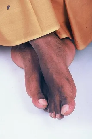 Close-up photo of a pair of bare feet crossed at the ankles, emerging from the hem of saffron-orange and pale ochre cloth. The feet rest on a plain white background. The toes and nails are clearly visible. No face or other body is shown.