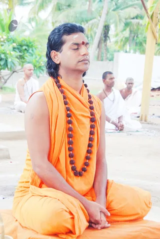 A man with shoulder-length swept-back dark hair sits cross-legged on an orange cushion with eyes closed and palms resting together in his lap, in meditation. He wears a sleeveless saffron/orange robe draped over one shoulder and a long necklace of rudraksha beads, with a red bindi and white vibhuti marks on his forehead. Behind him, three men in white ritual clothes sit in meditation on the sand under palm trees, one bald and one with a rudraksha mala. A plain outdoor setting with palms and a yellow pillar on the right suggests an open-air ashram gathering. a person cues: swept-back hair, saffron robe and rudraksha mala while meditating in the front position.