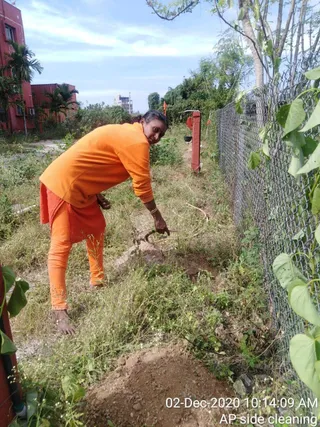 A woman in an orange long-sleeved top and orange leggings is bent over a dirt patch in an overgrown garden, using a tool to dig or clear soil beside a chain-link fence with climbing vines. A red-painted building, palm trees and a tall tree stand in the background under a partly cloudy sky. A timestamp in the corner reads 02-Dec-2020 10:14:09 AM and the label AP side cleaning. She is barefoot and wears a bangle on her wrist.