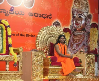 A seated man in saffron-orange robes with a rudraksha mala and a red bindi sits on an ornate gold throne with red cushions, smiling and holding his hands together in his lap. He is barefoot. Behind him a large orange backdrop shows a golden-toned deity murti with a crown, pearl and bead necklaces, and large earrings, flanked by red and yellow flower garlands. A microphone stands at his side, and a tray of offerings sits near the throne base. Kannada text is printed on the backdrop. a person: the saffron robes, rudraksha mala and ornate throne are the qualifying cues for the seated speaker.