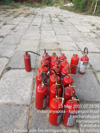 A photograph of red fire extinguishers lined up on a grey stone-paved path outdoors, with grass and weeds between the slabs and greenery and a red structure in the background. About sixteen extinguishers stand in rows, most with black hoses and yellow tags, labelled with POWDER and SAFE markings. A timestamp overlay reads 25 May 2021 07:28:36 and the location Vidyaranyapura - Nanjangud Road, Kenchanakuppe, Ramanagara, Karnataka. The caption reads Female side fire extinguisher going for refilling. No people are clearly visible apart from a distant figure at the top of the path, and no a person candidate appears.