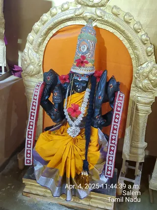 A black stone deity murti seated in a golden arched niche against an orange backdrop, shown in a temple shrine. The four-armed figure wears a tall jewelled crown decorated with red hibiscus flowers, a yellow-orange silk sari with a lavender and silver border, and a white flower garland with a pendant. A long black braid hangs down the front, and red-and-silver tasselled bands drape at the sides. The photo bears a timestamp reading Apr. 4, 2025 11:44:39 a.m., Salem, Tamil Nadu. No people are visible.
