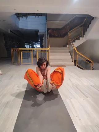 A young person with shoulder-length dark hair sits on a grey yoga mat on a pale marble floor, performing a seated forward-bend yoga pose and gripping their feet with both hands. They wear bright orange loose trousers with yellow-and-maroon trim, a bare upper body, and a brown beaded necklace, with white socks on their feet. Behind them are a golden-railed staircase and an open lobby with pale walls and recessed ceiling lights. The image is a photo of a single person.